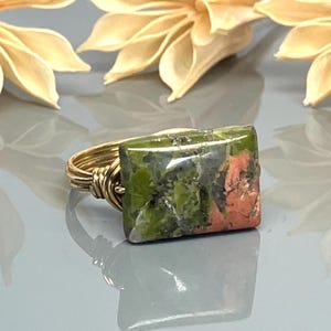 Peut inclure: Bague en fil d'or avec une pierre d'unakite rectangulaire. La pierre présente des tons verts, roses et gris. La bague est posée sur une surface réfléchissante avec des détails floraux flous.