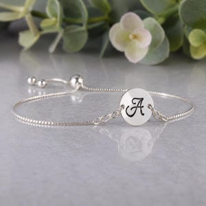 May include: A silver-toned adjustable bracelet featuring a delicate box chain and a circular charm engraved with the letter A in a black script font. The bracelet includes a sliding bead mechanism to adjust the fit, creating a sleek and simple design.