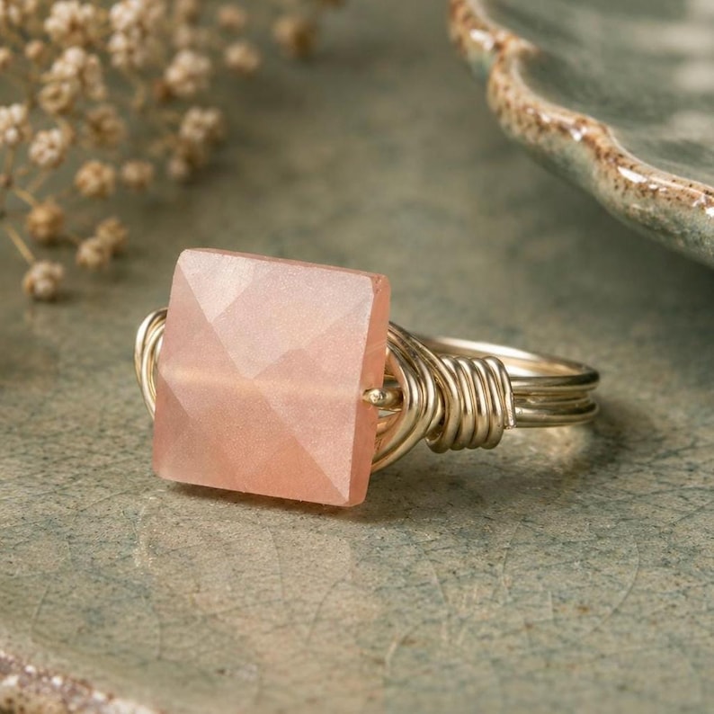 May include: A handcrafted ring featuring a square, faceted, peach-colored gemstone. The stone is set in a gold-colored wire wrap design. The ring is displayed on a textured, green surface, showcasing the jewelry's delicate details.