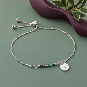 May include: A silver adjustable slider bracelet featuring a delicate box chain, a series of small silver and turquoise beads, and a round silver charm engraved with the capital letter I. The bracelet includes two small silver beads at the ends.