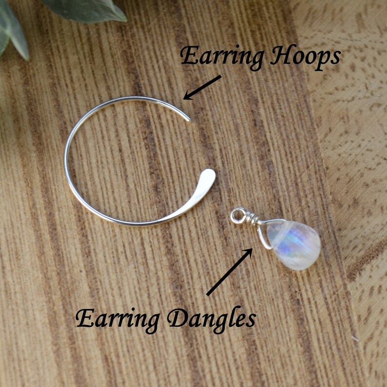 Pode incluir: Brincos de argola de prata com um pingente de pedra da lua. Os brincos de argola s&atilde;o abertos e curvos. O pingente de pedra da lua &eacute; enrolado em arame e tem uma apar&ecirc;ncia perolada. O texto na imagem diz "Earring Hoops" e "Earring Dangles".