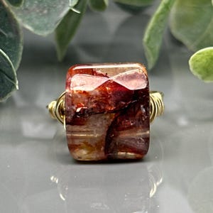 Peut inclure: Une bague avec une pierre précieuse carrée et une monture en fil doré. La pierre est un mélange de couleurs rouge, marron et transparent. La bague est posée sur une surface grise avec un feuillage vert en arrière-plan.