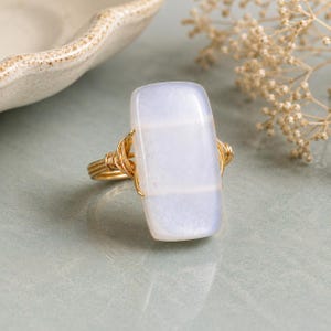 May include: A ring featuring a smooth, rectangular, pale blue chalcedony stone set in a gold-colored wire-wrapped band. The wire is coiled tightly around the sides of the stone, securing it to the thin, metallic band that encircles the finger.