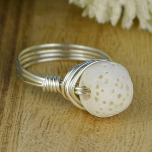 Peut inclure: Bague en fil d'argent avec une perle en pierre poreuse de couleur crème. L'anneau est composé de plusieurs brins de fil, et la perle est solidement enveloppée de fil. La bague est posée sur une surface en bois.