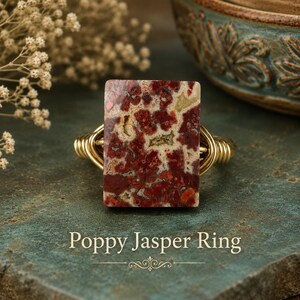 May include: A wire-wrapped ring featuring a rectangular poppy jasper stone with deep red, cream, and tan mottled patterns. The band is constructed from polished gold-toned wire coiled around the sides. The text Poppy Jasper Ring is visible.