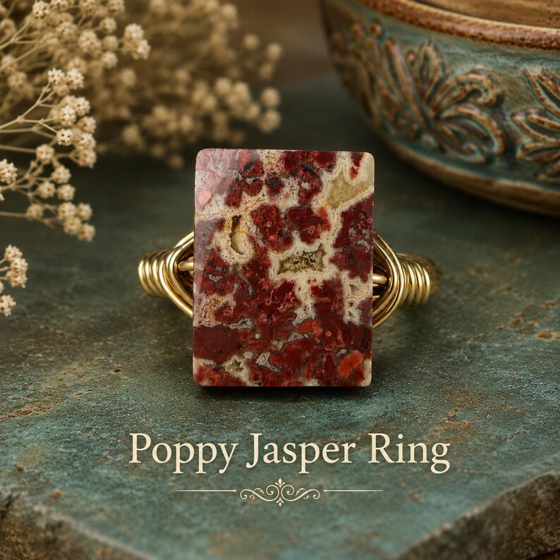 May include: A wire-wrapped ring featuring a rectangular poppy jasper stone with deep red, cream, and tan mottled patterns. The band is constructed from polished gold-toned wire coiled around the sides. The text Poppy Jasper Ring is visible.