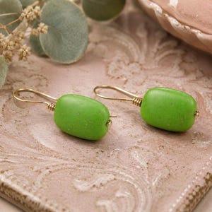 May include: A pair of green, oblong earrings with gold-colored wire hooks. The earrings have a smooth, matte finish and are displayed on a textured, pink surface. The earrings are a simple, yet elegant accessory.