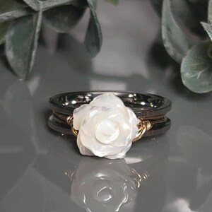 A white carved rose/flower ring with a two tone gold and stainless steel spinner ring band is displayed on a reflective surface, surrounded by green leaves.