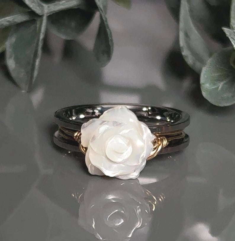 A white carved rose/flower ring with a two tone gold and stainless steel spinner ring band is displayed on a reflective surface, surrounded by green leaves.