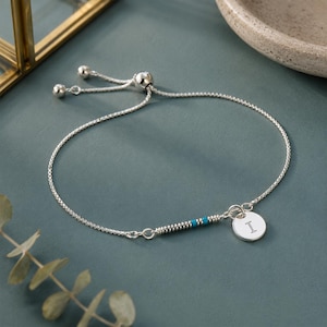 May include: A silver adjustable bracelet featuring a delicate box chain, a small circular charm engraved with the letter I, and a decorative segment of silver beads interspersed with two bright turquoise blue beads near the charm.