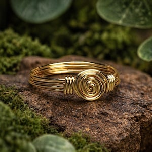 May include: A gold-colored ring with a spiral design at the center. The ring is made of multiple strands of wire, wrapped to form the band and the central spiral. The ring is displayed on a textured, brown stone, with green moss in the background.