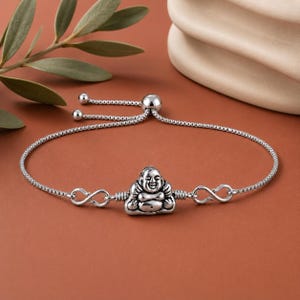 May include: A silver-toned bracelet featuring a central charm of a smiling, seated figure with a rounded belly. The charm is flanked by two infinity-shaped links on a thin box chain, which includes an adjustable sliding bead closure with two small spheres.