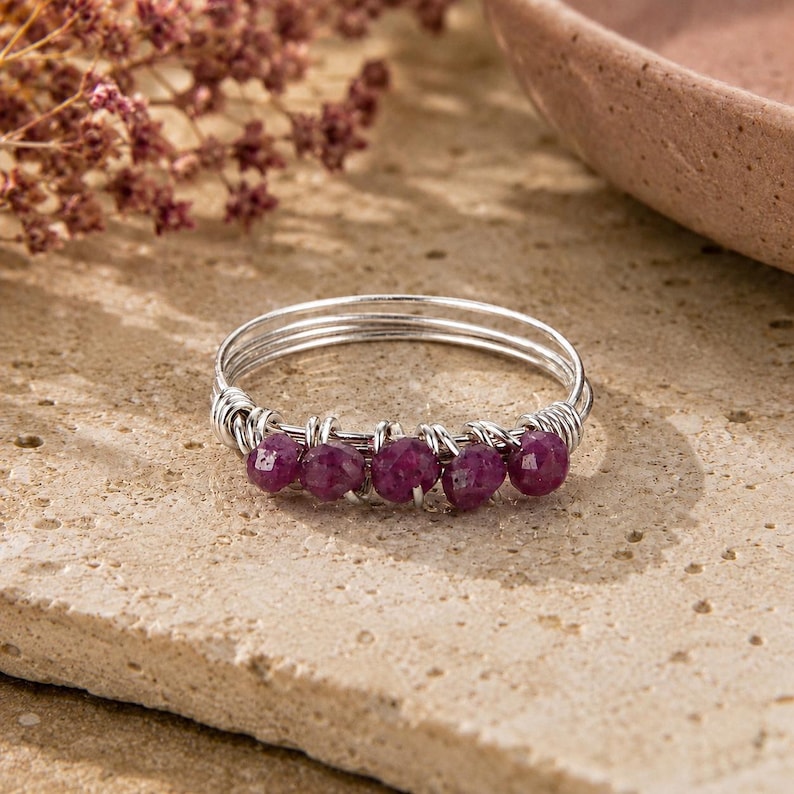 May include: A silver-toned wire-wrapped ring featuring five small, faceted, deep magenta-colored gemstone beads arranged in a row along the front of the band. The band is constructed from multiple thin strands of silver-colored wire coiled together.