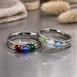 May include: Two silver rings with colorful beaded accents. One ring features red, blue, and green beads, while the other has green, light blue, and blue beads. The beads are set in a wire-wrapped design, adding a unique touch to the rings.