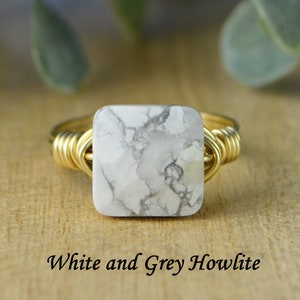 May include: A gold-toned wire-wrapped ring featuring a square, white and gray howlite stone. The stone has a marbled appearance. The text "White and Grey Howlite" is displayed below the ring.