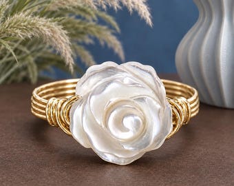May include: A gold-toned wire cuff bracelet featuring a central white mother-of-pearl rose ornament. The band is constructed from multiple parallel strands of gold-coloured wire that wrap around the base of the carved floral centerpiece.