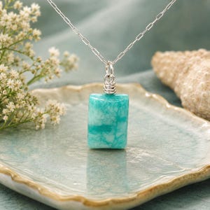 May include: A rectangular pendant necklace with a silver chain. The pendant is a vibrant turquoise color with white veining. The pendant is approximately 1 inch long and is displayed on a light blue ceramic plate.