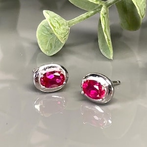 May include: A pair of silver stud earrings with oval-shaped ruby gemstones.