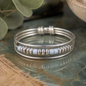 May include: A silver bangle bracelet with three parallel bands. The bracelet features a row of small, light blue beads and silver beads. The bracelet is displayed on a reflective, green surface.