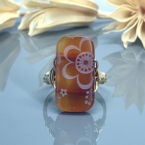 Peut inclure: Une bague en fil d'or avec une pierre rectangulaire en verre orange ornée d'un motif floral blanc.