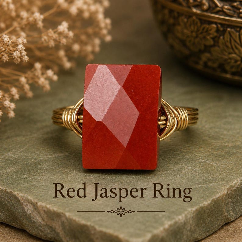 May include: A ring featuring a rectangular, faceted red jasper gemstone set on a gold-toned wire-wrapped band. The text Red Jasper Ring is printed below the jewelry piece. The stone has a deep red color with a geometric, multi-faceted surface.