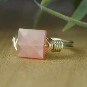 May include: A silver-toned ring featuring a square, faceted, peach-colored gemstone. The gemstone is the focal point, set in a wire-wrapped design. The ring is displayed on a wooden surface, with a blurred green plant in the background.