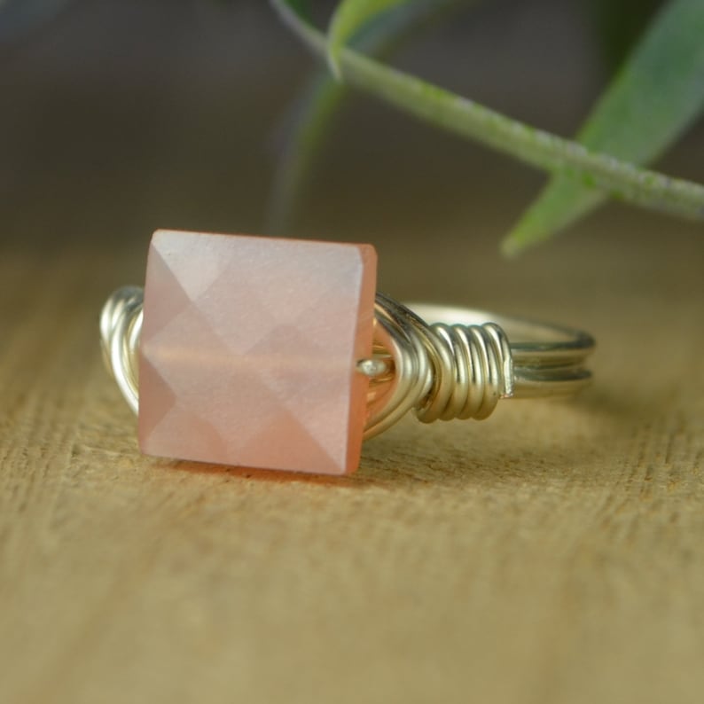 May include: A silver-toned ring featuring a square, faceted, peach-colored gemstone. The gemstone is the focal point, set in a wire-wrapped design. The ring is displayed on a wooden surface, with a blurred green plant in the background.