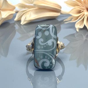 Peut inclure: Une bague en fil d'or avec une pierre rectangulaire à motifs verts et blancs. La pierre présente un motif de tourbillon blanc sur un fond vert.