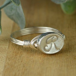 May include: A silver ring with a wire-wrapped band and a round silver charm with the letter "R" engraved on it.