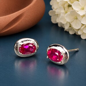 May include: A pair of silver stud earrings featuring oval-cut, vibrant pinkish-red gemstones set in polished metal frames. Each earring has a secure post backing and a smooth, reflective surface surrounding the central faceted stone.