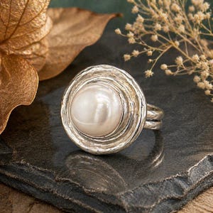 May include: A silver ring with a large, lustrous pearl center. The ring's setting has a textured, swirling design around the pearl. The ring is displayed on a dark, textured surface, with a blurred background of dried leaves and flowers.
