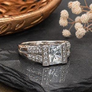 May include: A silver engagement ring with a large, clear, rectangular center stone. The band is adorned with smaller, sparkling stones. The ring is displayed on a dark, textured surface, reflecting the light.