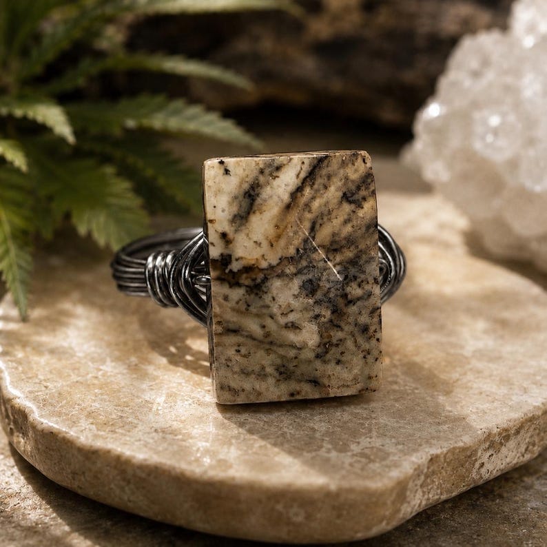 May include: A ring featuring a rectangular, polished stone with a cream, tan, and dark brown marbled pattern. The stone is secured by a dark, metallic wire-wrapped band that coils around the sides to hold the stone in a vertical orientation.