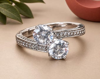 May include: A bypass ring crafted from silver-toned metal featuring two prominent round-cut clear stones set side-by-side. The band is split into two sections, each encrusted with a row of small, sparkling clear stones along the outer edges.