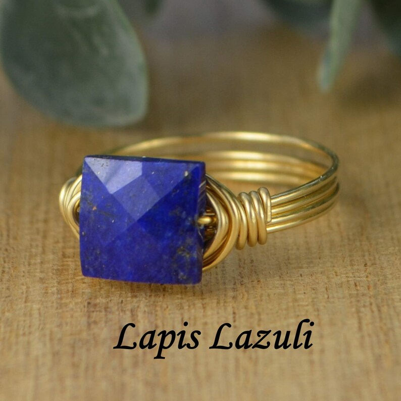 May include: A gold-toned wire-wrapped ring featuring a square, faceted, deep blue lapis lazuli stone. The ring is set against a wooden surface, with the text "Lapis Lazuli" below the ring.