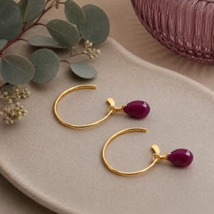 May include: A pair of gold hoop earrings with a teardrop-shaped, deep red gemstone pendant. The earrings are displayed on a neutral-toned surface with a sprig of greenery in the background. The gemstones have a faceted cut.