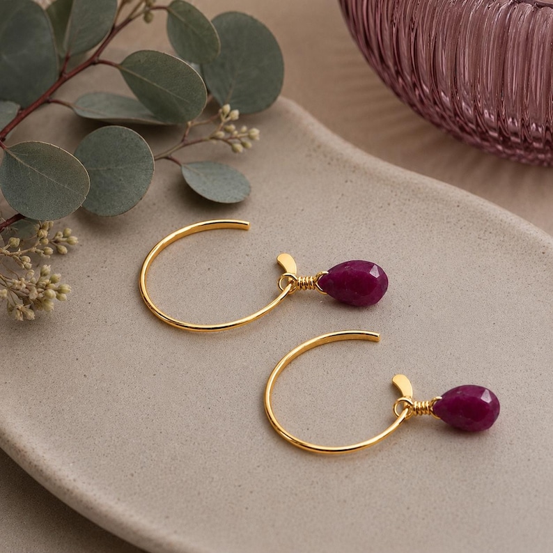 Pode incluir: Um par de brincos de argola dourados com um pendente de pedra preciosa vermelha escura em forma de gota. Os brincos s&atilde;o exibidos em uma superf&iacute;cie de tom neutro com um galho de vegeta&ccedil;&atilde;o ao fundo. As pedras preciosas s&atilde;o facetadas.