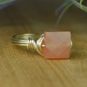 May include: A handmade ring with a square, peach-colored gemstone. The gemstone is set in a silver wire wrap design. The ring is displayed on a wooden surface, showcasing the jewelry's craftsmanship and color.