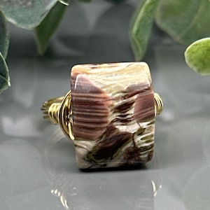 May include: A rectangular gemstone ring with a gold-colored wire band. The stone features a mix of brown, cream, and green hues with a layered pattern. The ring is set against a blurred background with greenery.