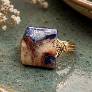 May include: A ring featuring a rectangular, polished stone with a marbled pattern of deep blue, cream, and reddish-brown tones. The stone is secured by a gold-colored wire-wrapped band that coils around the sides to hold the setting in place.