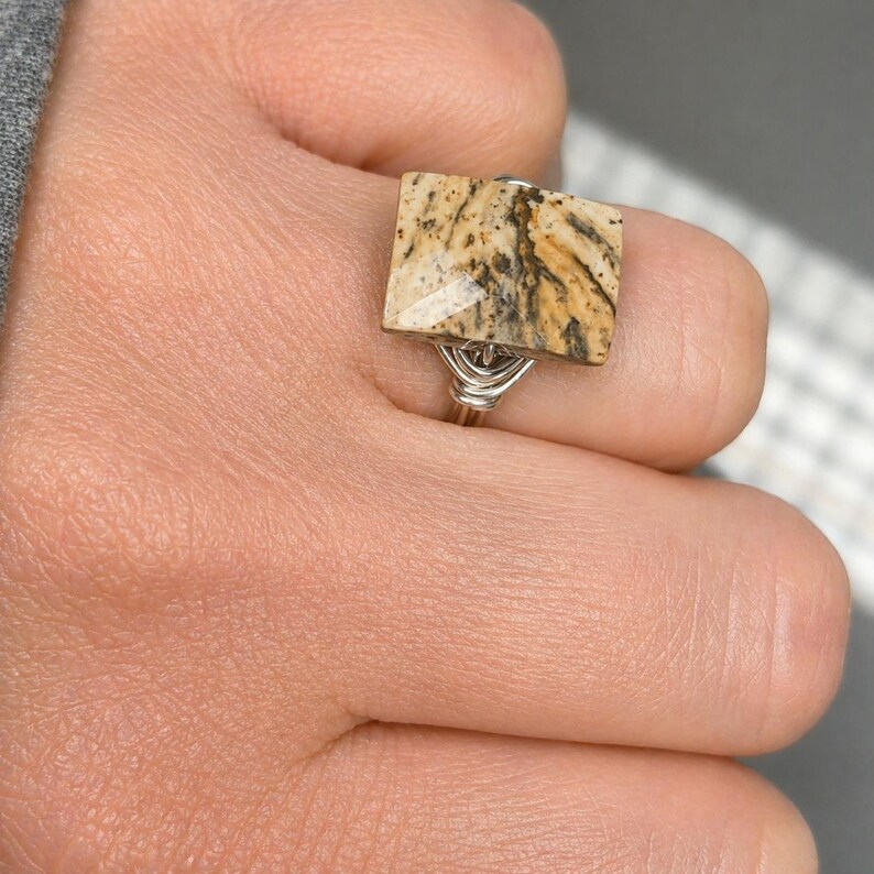 May include: A ring featuring a rectangular, tan and brown patterned stone set on a silver-colored wire-wrapped band. The stone displays natural dark veining and a smooth, polished surface, securely held in place by the coiled metal wire design.