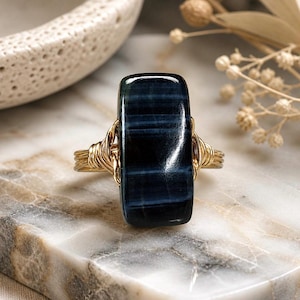 May include: A rectangular blue tiger eye stone ring with a gold wire wrap. The stone has dark blue and black stripes. The ring is set on a marble surface.