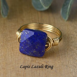 May include: A gold-toned wire-wrapped ring featuring a square, faceted lapis lazuli stone. The deep blue stone has flecks of gold. The ring is displayed on a wooden surface. Text on the image reads "Lapis Lazuli Ring."