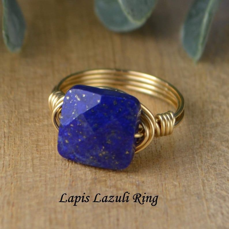 May include: A gold-toned wire-wrapped ring featuring a square, faceted lapis lazuli stone. The deep blue stone has flecks of gold. The ring is displayed on a wooden surface. Text on the image reads "Lapis Lazuli Ring."