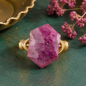 May include: A ring featuring a large, faceted, hexagonal-shaped magenta gemstone set in a gold-colored wire-wrapped band. The stone displays deep pink and purple hues with natural inclusions, secured by multiple loops of thin, metallic gold wire.