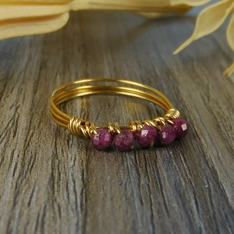May include: A gold-toned wire ring featuring five small, faceted, deep magenta-colored beads. The beads are secured along the front of the band with thin gold-toned wire wraps, creating a delicate, layered appearance for this piece of jewelry.