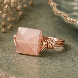 May include: A handcrafted ring featuring a square, faceted, light pink gemstone. The gemstone is set in a copper-colored wire wrap design. The ring is displayed on a textured, green surface, showcasing the jewelry's unique craftsmanship.