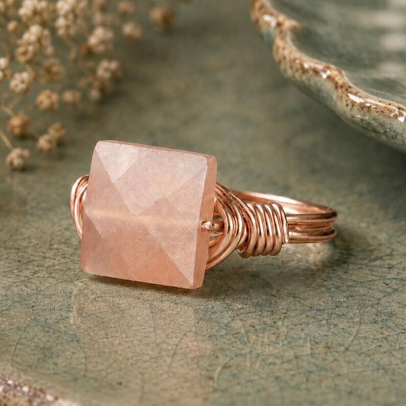 May include: A handcrafted ring featuring a square, faceted, light pink gemstone. The gemstone is set in a copper-colored wire wrap design. The ring is displayed on a textured, green surface, showcasing the jewelry's unique craftsmanship.