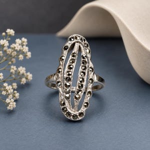 May include: A silver ring featuring an elongated oval design with a central vertical opening. The metal frame is textured and encrusted with numerous small, dark, faceted marcasite stones arranged in rows along the outer and inner edges.