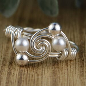 May include: A silver-toned wire ring with a spiral design, featuring three pearl-like beads and two silver beads. The ring is set against a blurred wooden background, highlighting the intricate wirework and the contrast of the shiny beads.